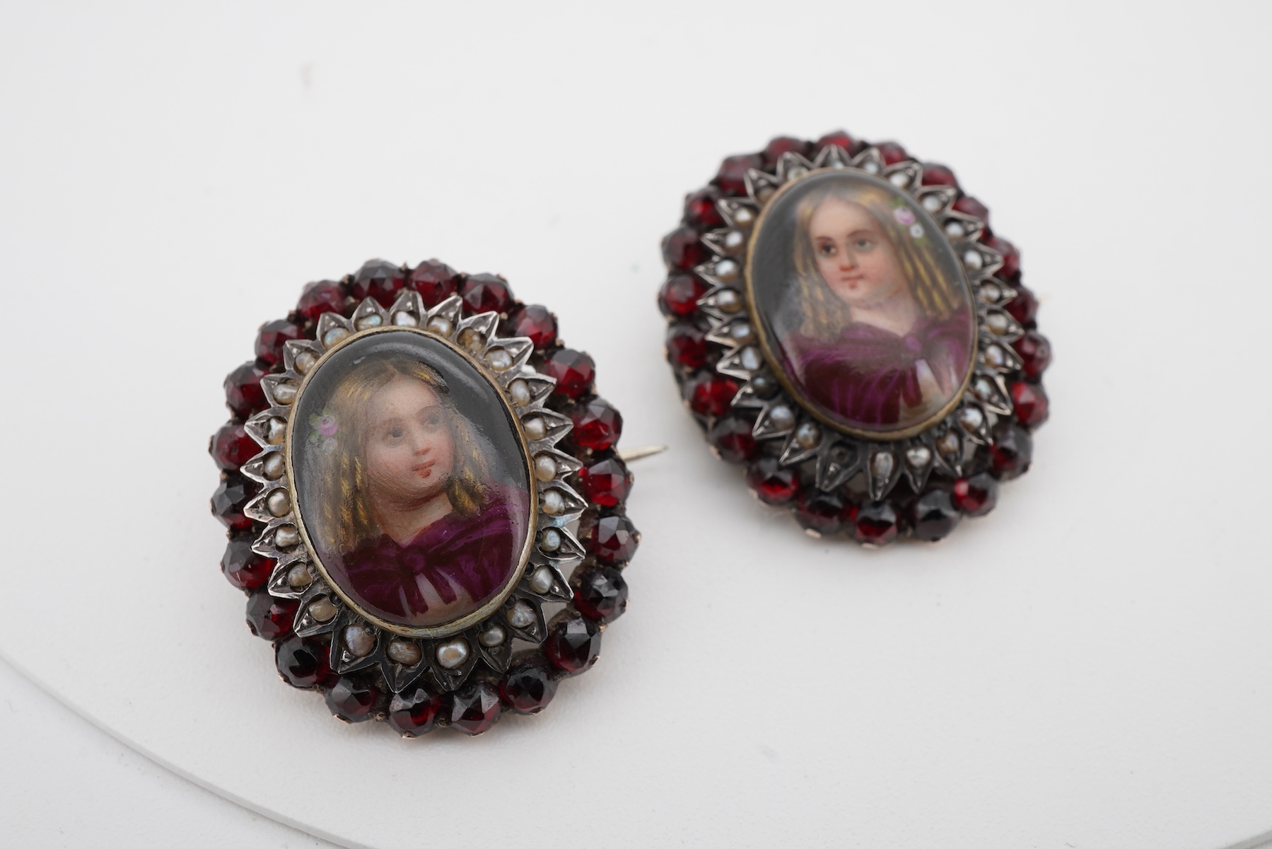 A pair of early 20th century yellow metal mounted brooches, with central oval panel painted with the bust of a lady and bordered by concentric rows of seed pearls and garnets (adapted), 24mm by 20mm.
